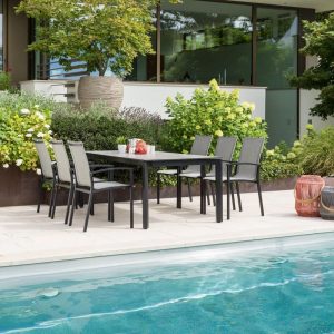 Ein Tisch mit sechs Stühlen steht zwischen dem Garten vor einer breiten Glasfront eines Hauses und einem Swimmingpool